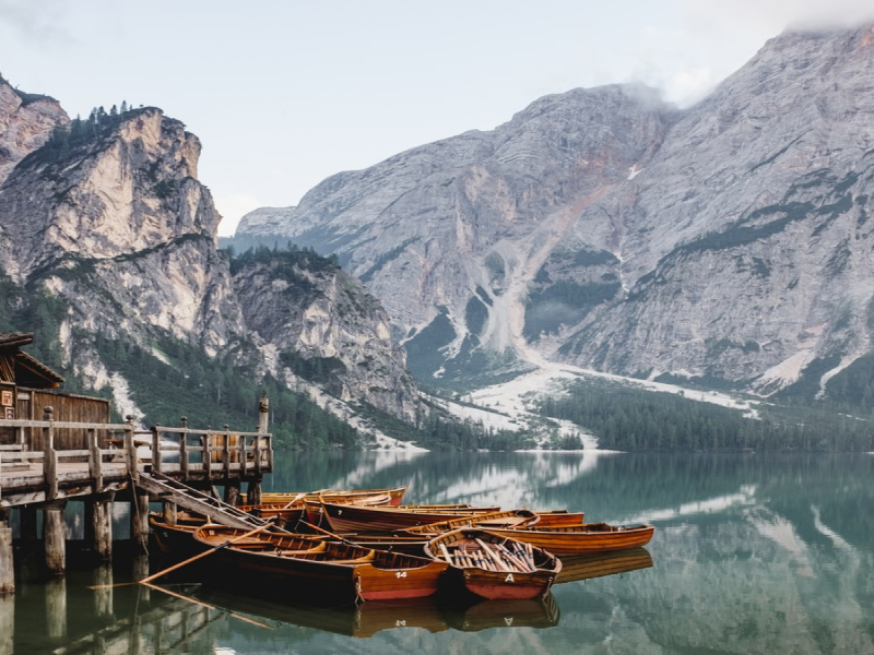Wooden cabins rest beside a quiet dock while vintage boats float calmly on a mirrored lake. Surrounded by rugged peaks and pine trees, the place evokes peace, tradition, and natural beauty. The perfect reflection in the water creates symmetry and stillness. An ideal hideaway to unplug and enjoy serenity.