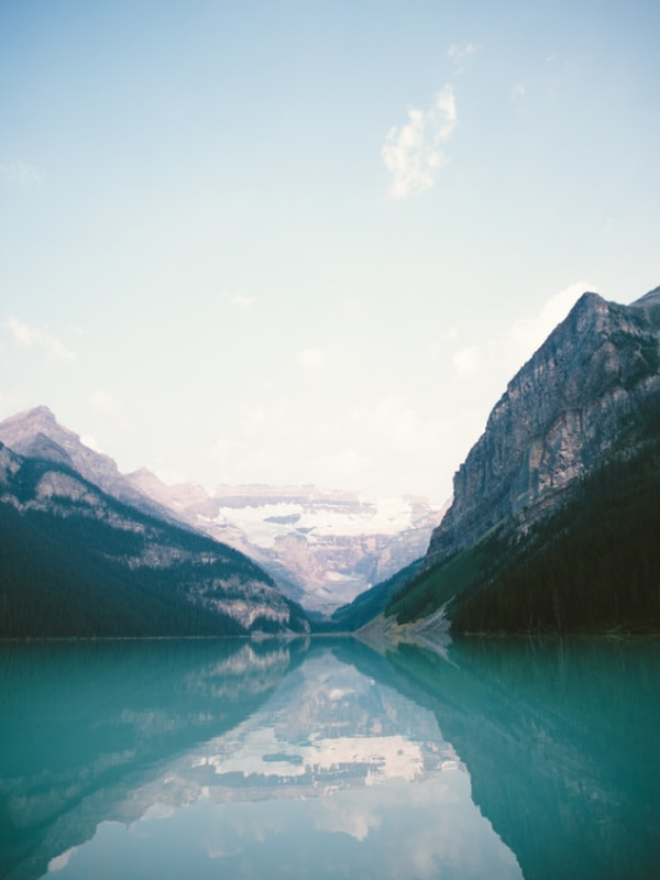 A turquoise lake reflects snow-covered peaks with stunning clarity. The stillness of the water creates a perfect natural mirror, doubling the magnitude of the landscape. With a clear sky and towering mountains, the scene feels open, fresh, and deeply contemplative. Pure, untouched nature at its most beautiful and calm.
