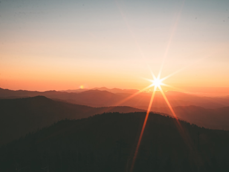 A warm sun sets on the horizon, casting shadows and golden light over distant mountains. The soft layering of the hills fades into the distance, evoking calm and closure. The scene glows with the perfect end of day, inviting quiet thought and gratitude for natural beauty.