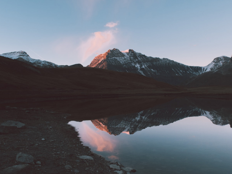 A snowy mountain is perfectly reflected in a calm lake at dawn. The sun’s pink light touches the summit, blending beautifully with the pale blue sky. This photo captures the precise moment between night and day—silent, majestic, and perfectly composed. Nature in balance and harmony.